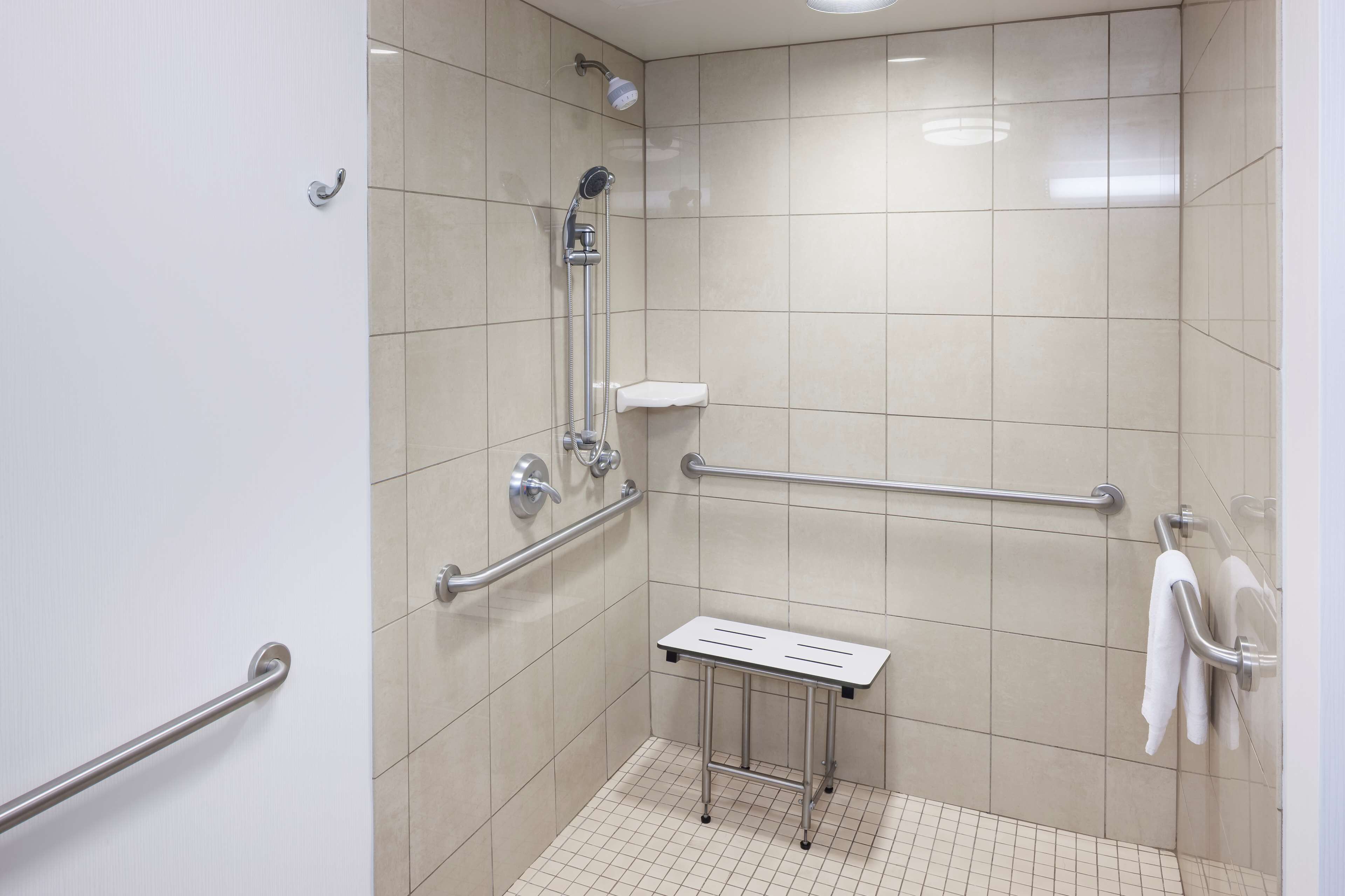 Un espacio de ducha pequeño con azulejos, barras de apoyo, un cabezal de ducha manual y un perchero blanco.