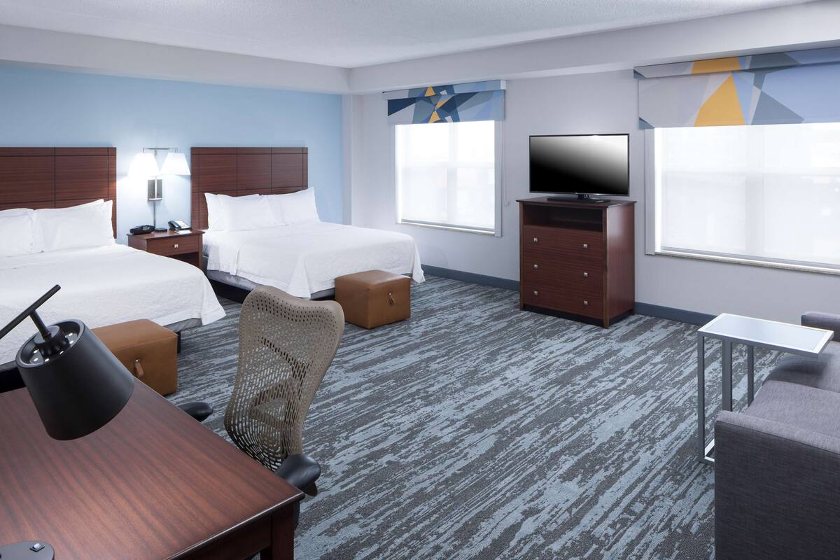 Ein Hotelzimmer mit zwei Betten, einem Schreibtisch, einem Stuhl, einem Fernseher, einem Schminktisch und einem Fenster mit Jalousien.