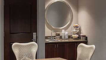 A hotel room with a wooden dining table, two wicker chairs, a round mirror, a sink, and a breakfast bar with various items.