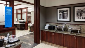 A hotel kitchen with a buffet setup, including a coffee station, condiment dispensers, and a sign that reads "TASTE POUR MIX LAUGH."