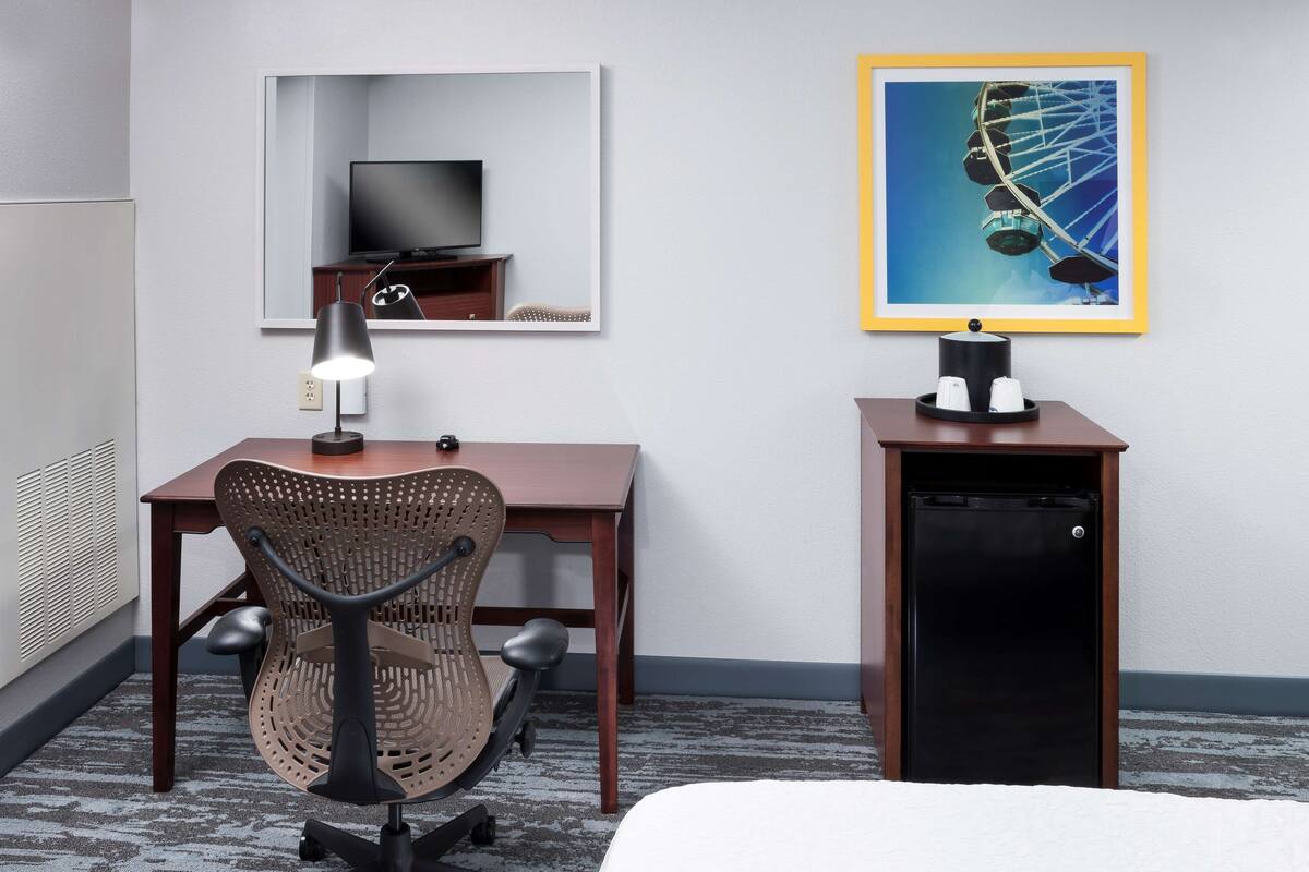 Ein Hotelzimmer mit Schreibtisch, Stuhl, Fernseher, Minikühlschrank und einem gerahmten Bild von einem Riesenrad.