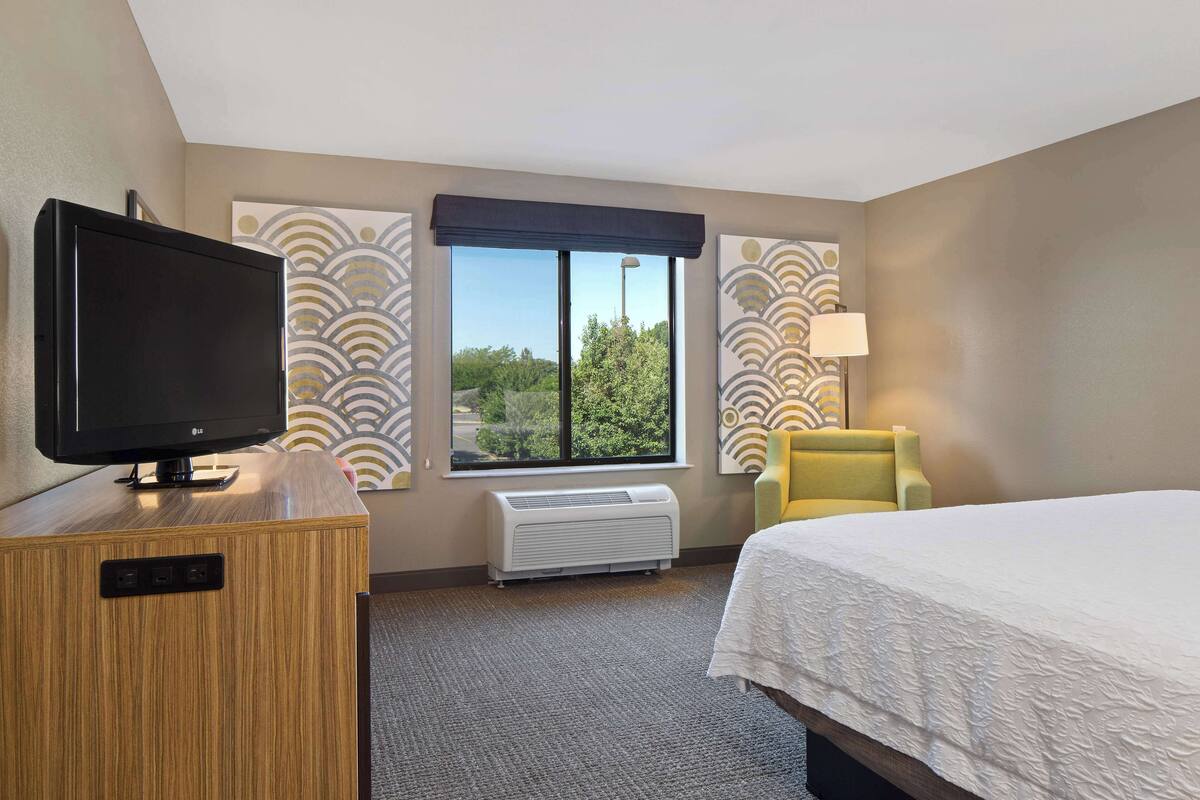 A hotel room with a bed, a television on a wooden dresser, a window with a view of greenery, and a yellow armchair.