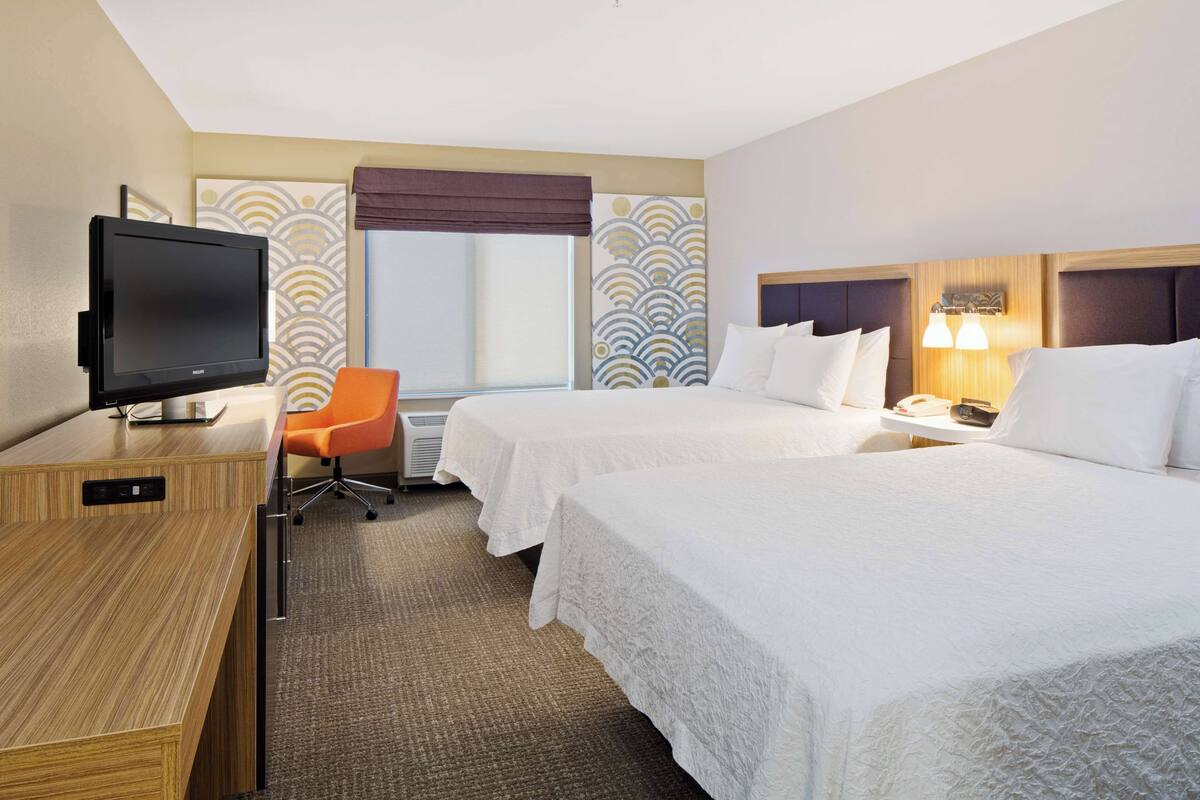 A hotel room with two beds, a desk with a flat-screen TV, an orange chair, and a window with a view.