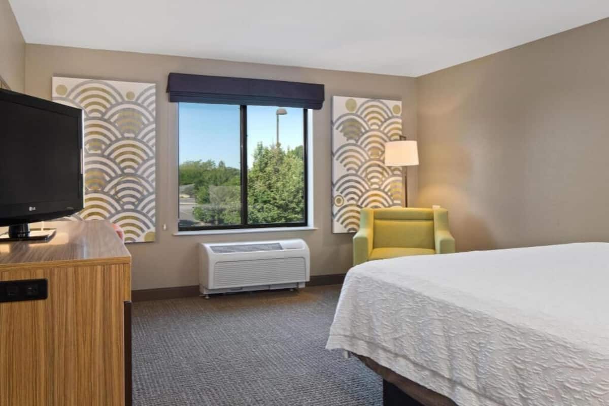 Habitación de hotel con cama, televisor, silla, ventana con vistas y pared decorada.