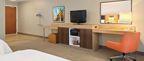 Habitación de hotel con cama, escritorio con computadora, televisor, lámpara y un cuadro en la pared.