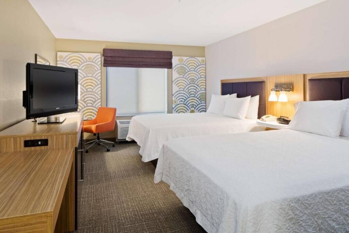 A hotel room with two beds, a desk with a flat-screen TV, an orange chair, a window with a view, and a wall with a decorative pattern.
