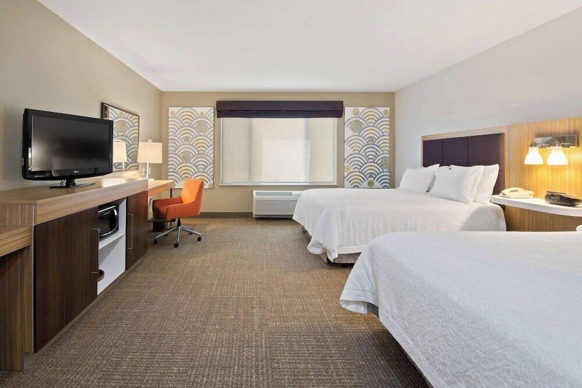 A hotel room with two beds, a desk with a TV, a chair, and a window with patterned curtains.
