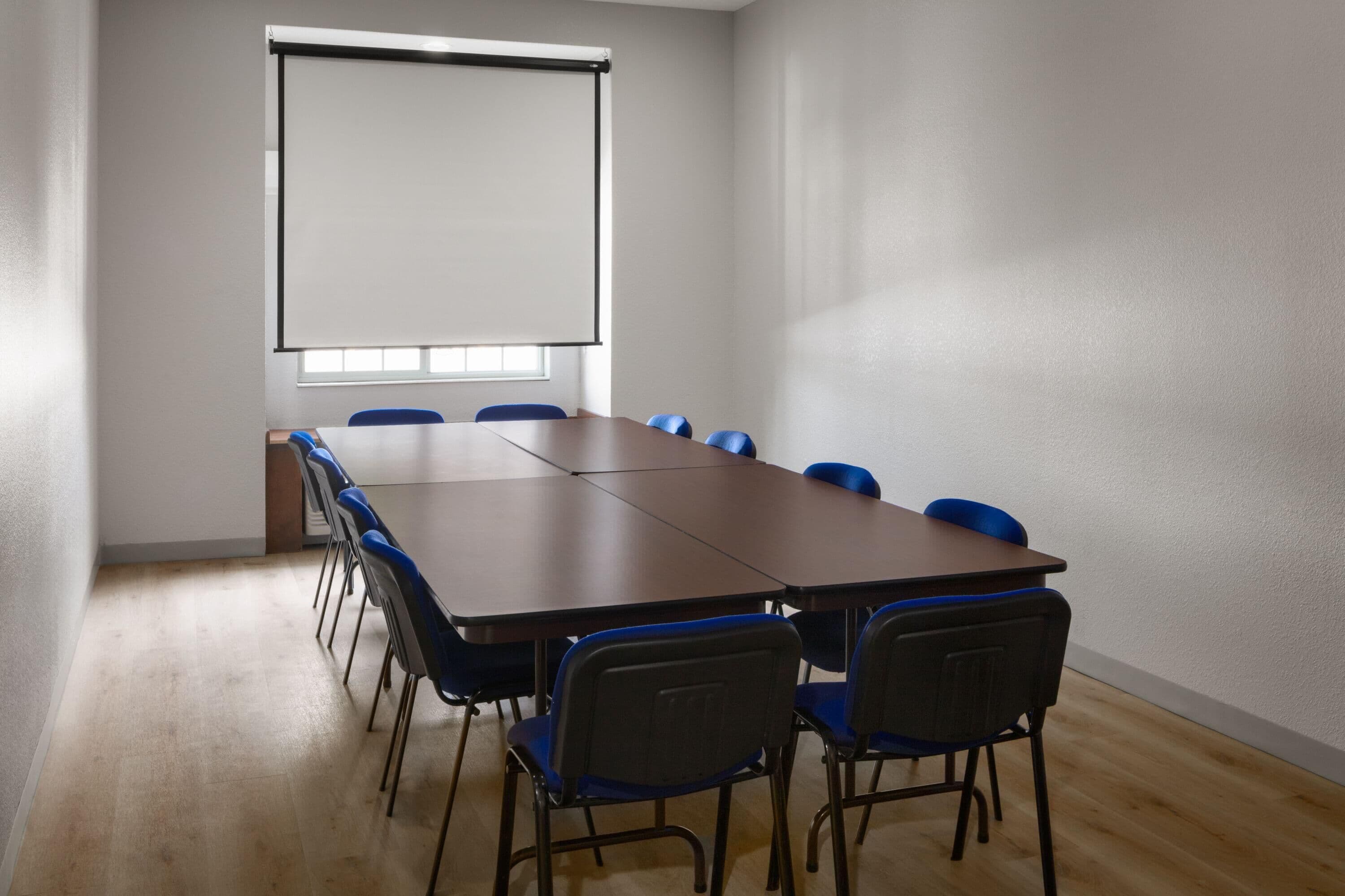 A conference room with a long table, blue chairs, a projector screen, and a window.