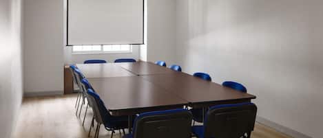A conference room with a long table, blue chairs, a projector screen, and a window.