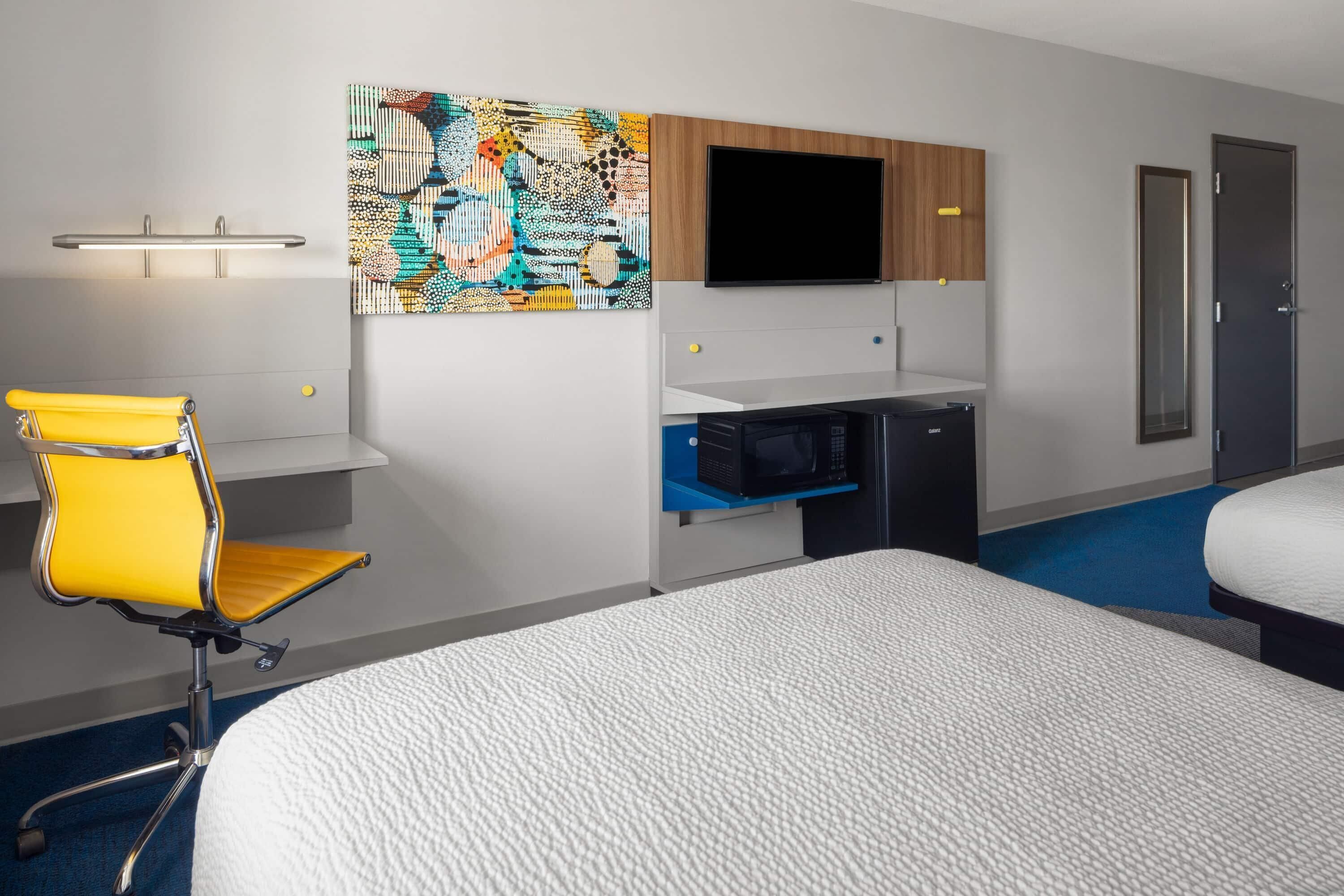 A hotel room with a bed, a desk with a computer, a TV, a bathroom, and a colorful artwork on the wall.