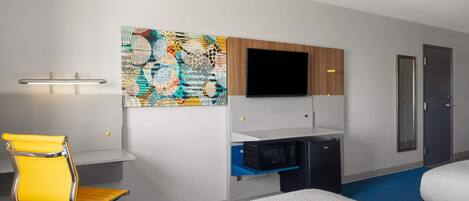 A hotel room with a bed, a desk with a computer, a TV, a bathroom, and a colorful artwork on the wall.