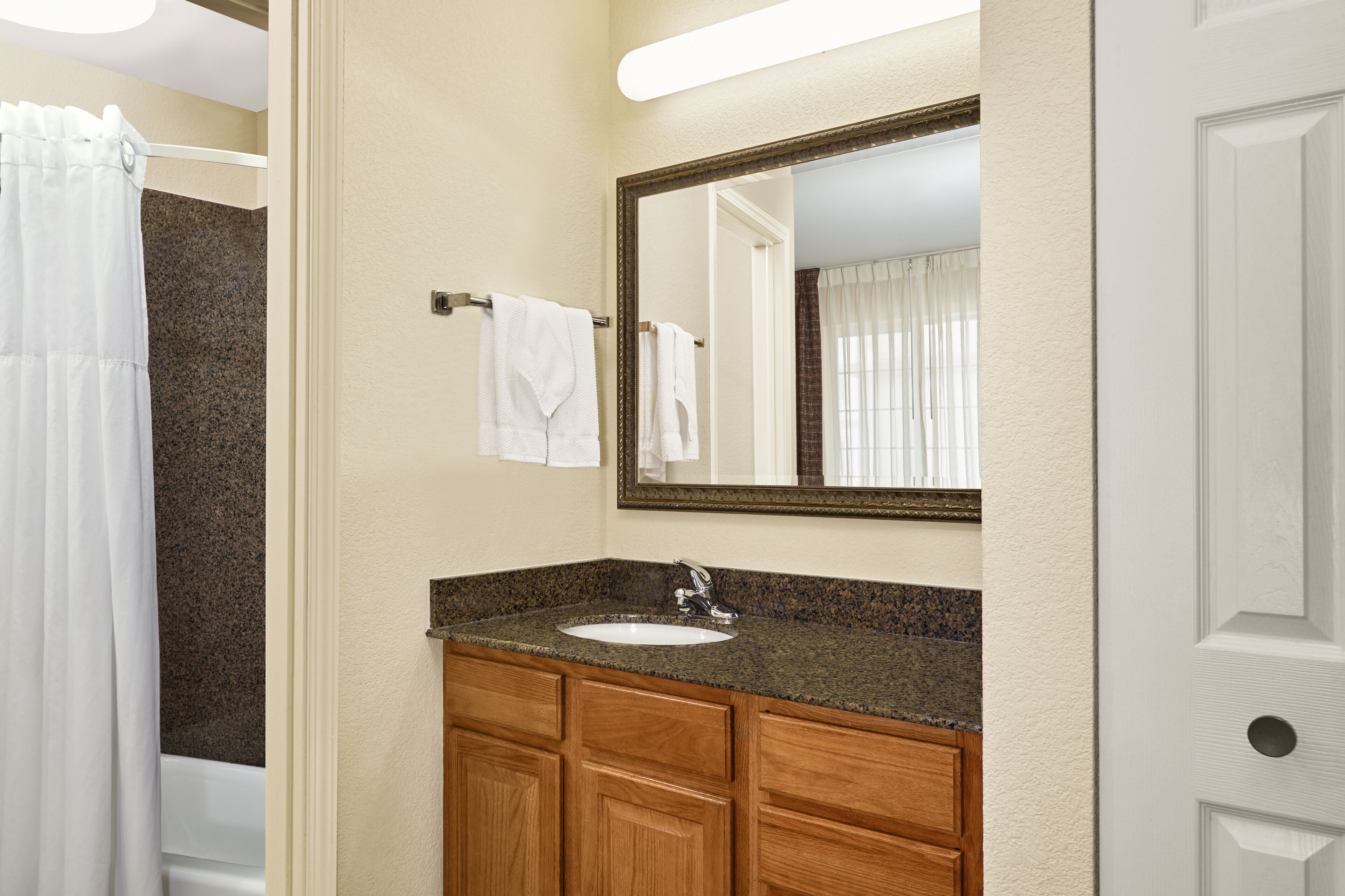A bathroom with a granite countertop sink, wooden cabinets, a large mirror, and a white shower curtain.