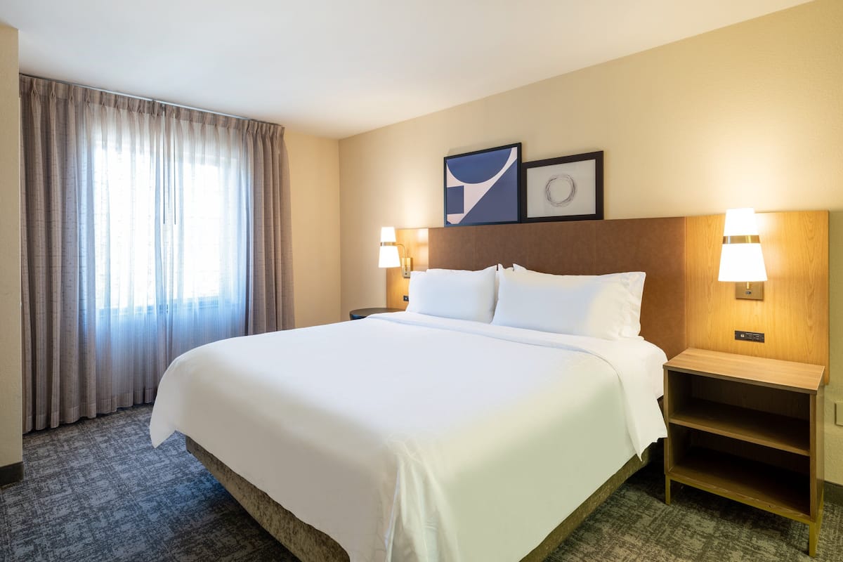 Une chambre d’hôtel avec un grand lit, deux tables de chevet avec des lampes, une tête de lit et une fenêtre avec des rideaux.