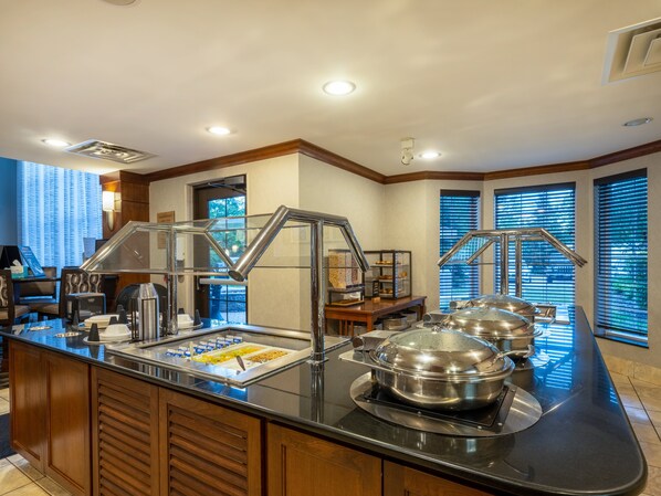 Un espace buffet délimité par une paroi en verre, avec des plats de service en acier inoxydable et un coin café.