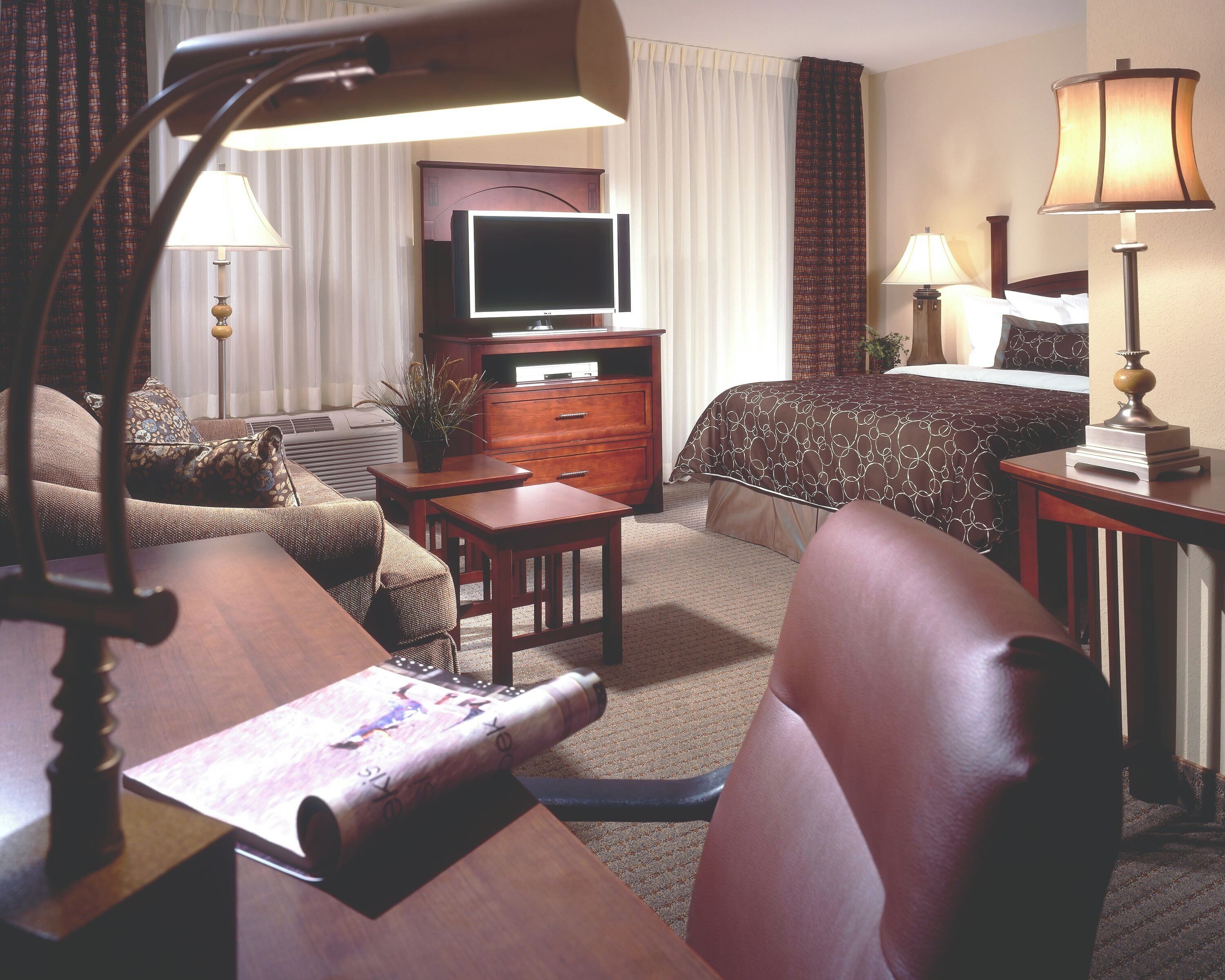 A hotel room with a bed, a desk, a television, a lamp, and a plant.