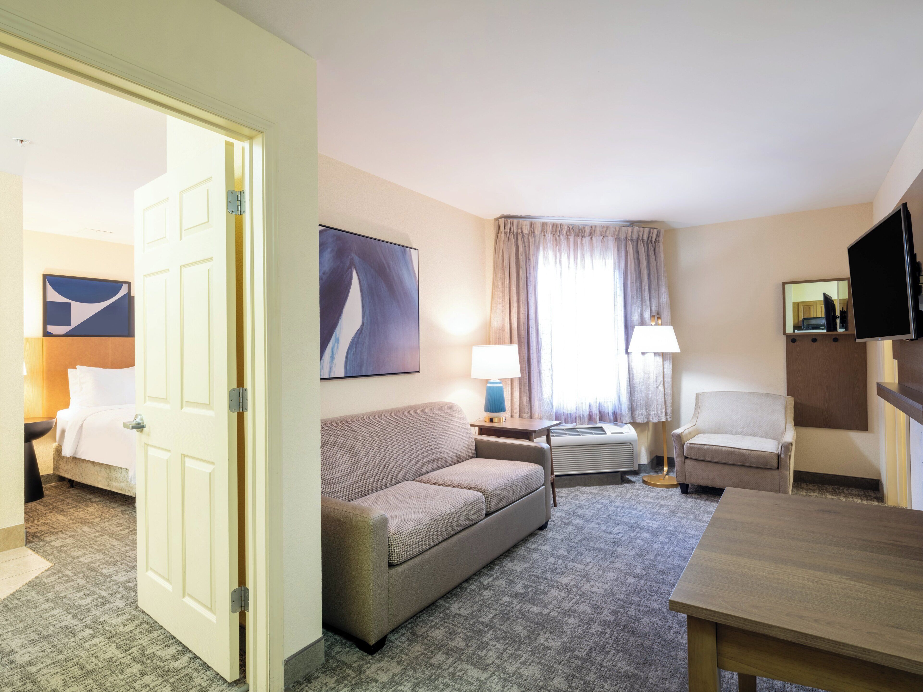 A hotel room with a bed, a sofa, a desk, a chair, a television, and a window with curtains.