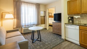 Ein Hotelzimmer mit einer Couch, einem kleinen runden Tisch, einem Fernseher, einer Küchenzeile und einem sichtbaren Badezimmer durch eine offene Tür.