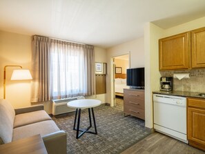Ein Hotelzimmer mit einer Couch, einem kleinen runden Tisch, einem Fernseher, einer Küchenzeile und einem sichtbaren Badezimmer durch eine offene Tür.