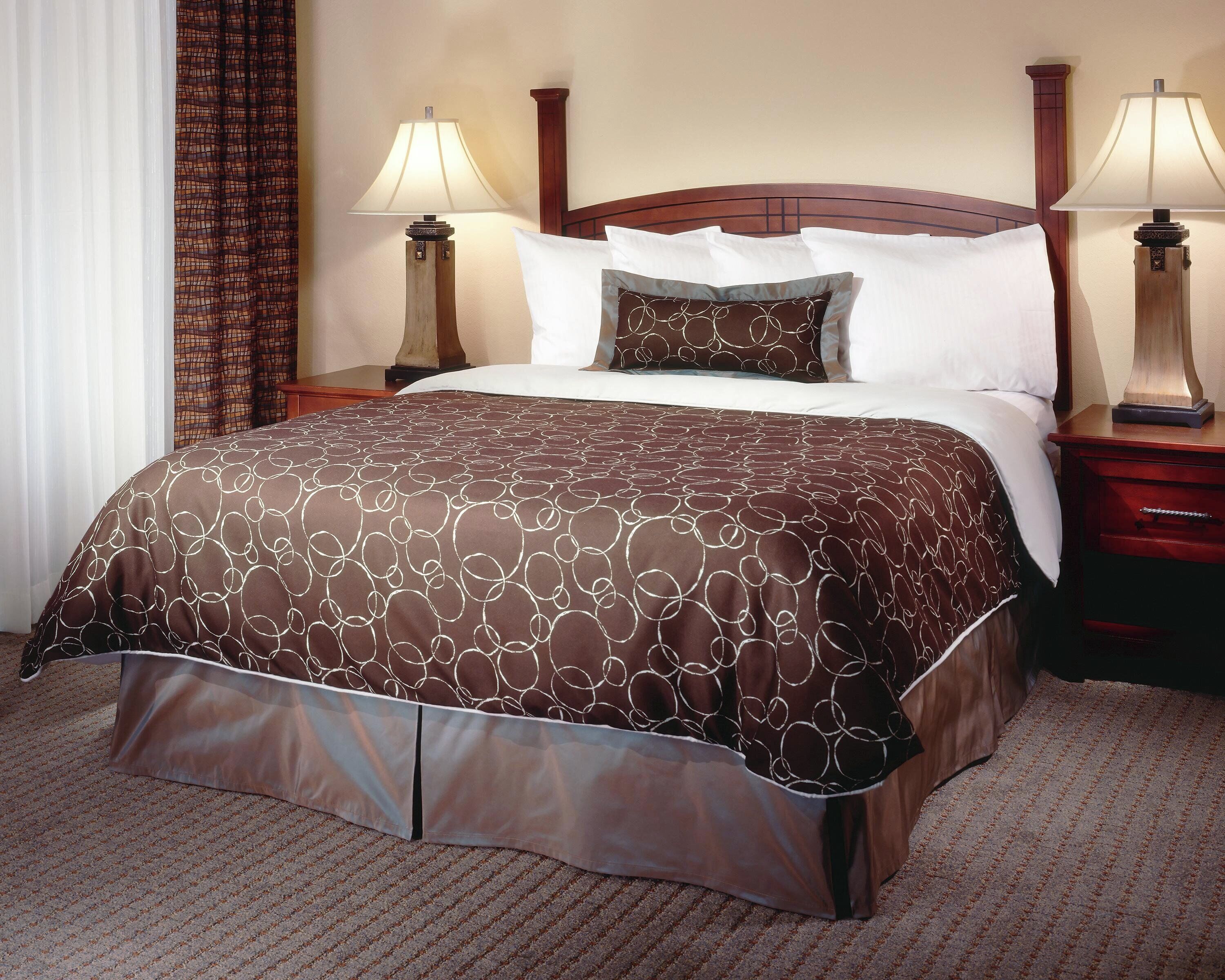 A hotel room with a large bed, two bedside lamps, a wooden headboard, and a patterned bedspread.