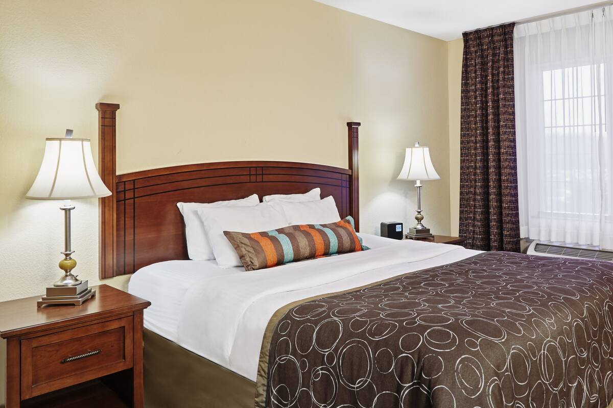 Une chambre d’hôtel avec un grand lit, deux tables de chevet, une petite table, une lampe, une fenêtre avec des rideaux et un climatiseur fixé au mur.