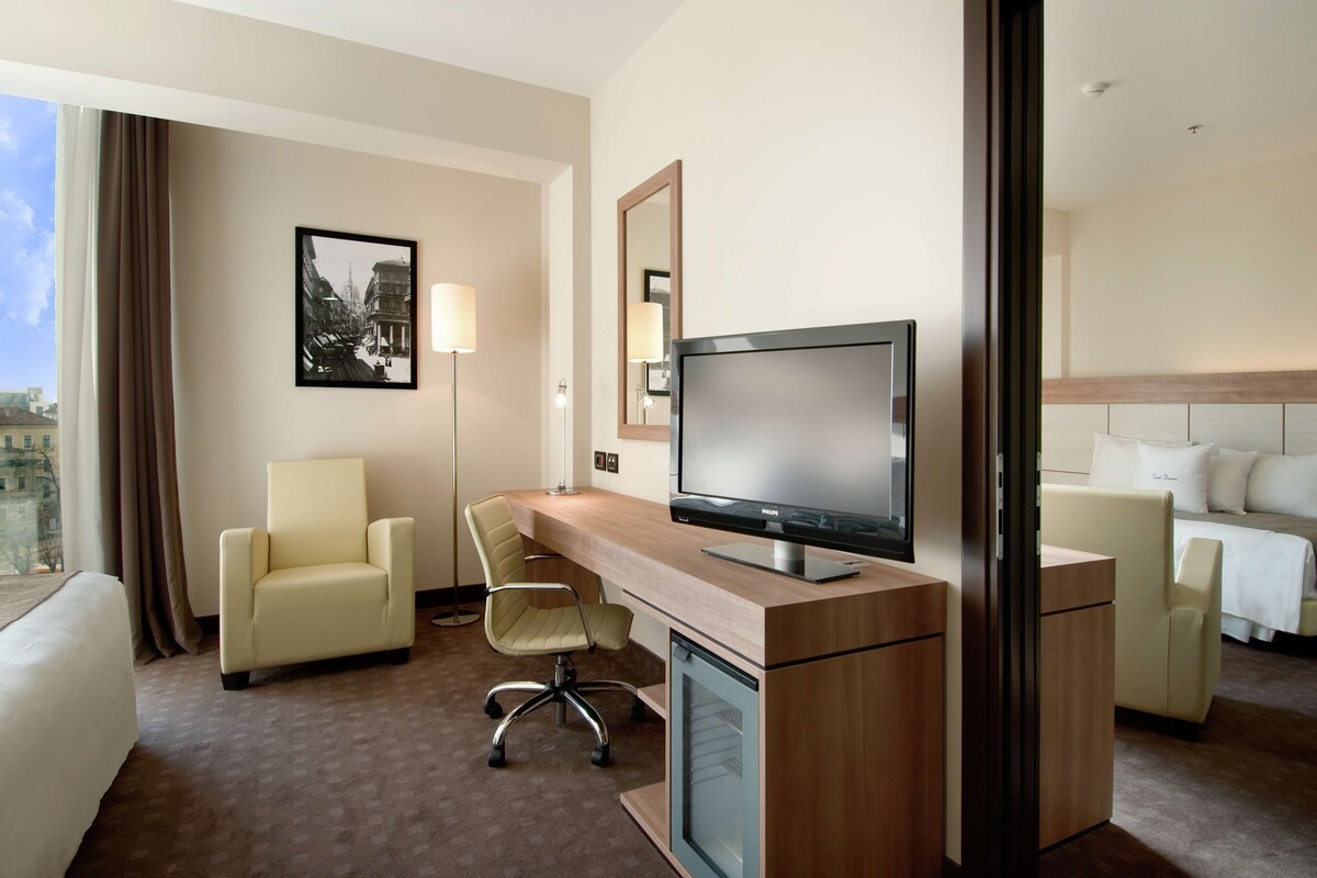 Une chambre d’hôtel équipée d’un bureau, de deux chaises, d’une télévision, d’un lit et d’une grande fenêtre avec des rideaux.