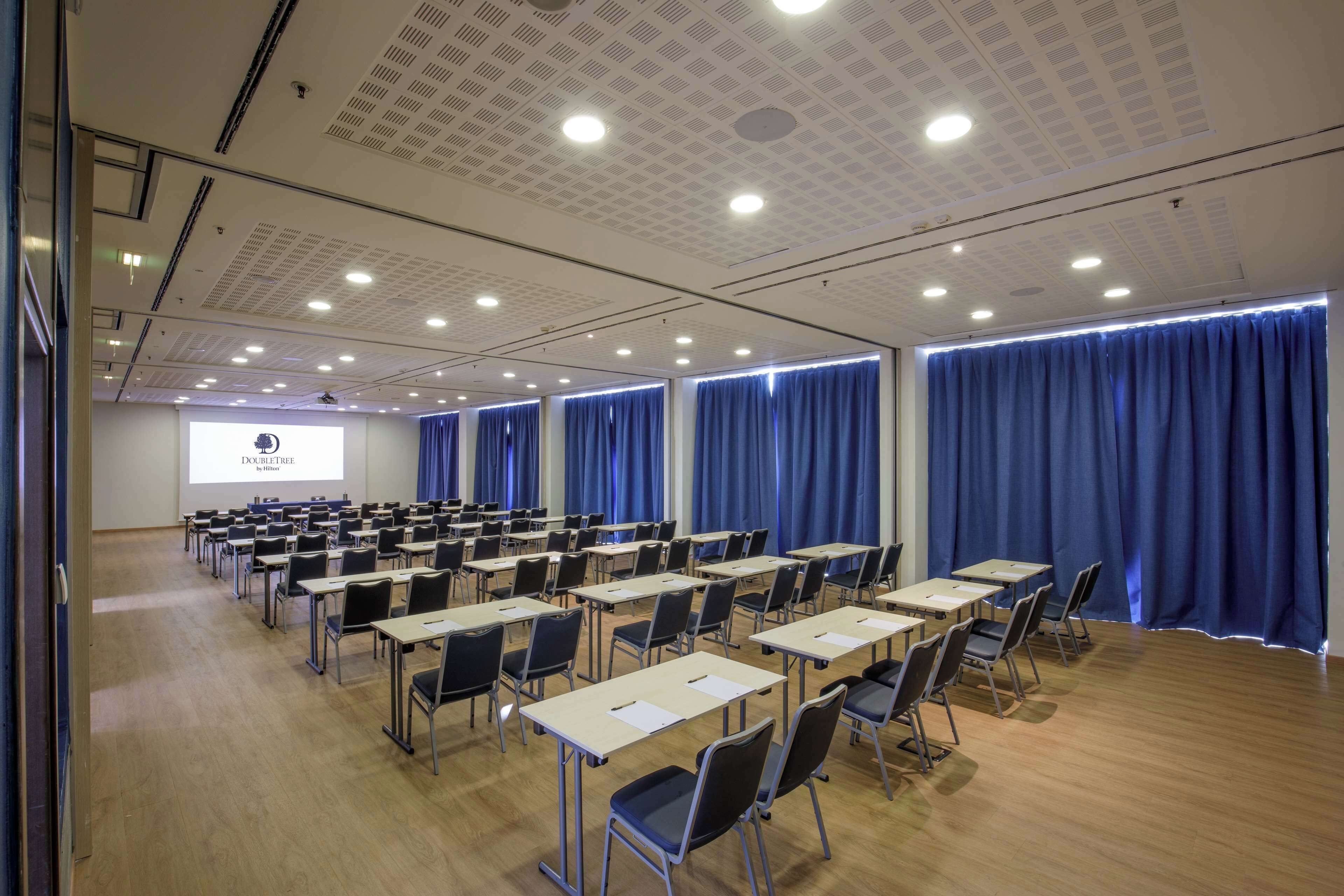 A conference room with rows of tables and chairs, a projector screen, and blue curtains.