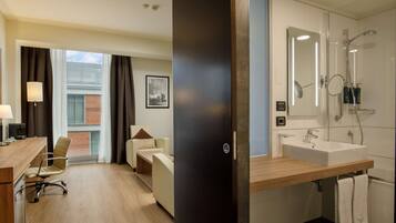 Une chambre d’hôtel avec un bureau en bois, une chaise, un lit, une grande fenêtre avec des rideaux, une salle de bain avec un lavabo et un miroir, et une douche.