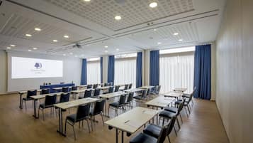 Konferenssitila, jossa on riveissä pöytiä ja tuoleja, projektorikangas ja suuret ikkunat, joissa on verhot.