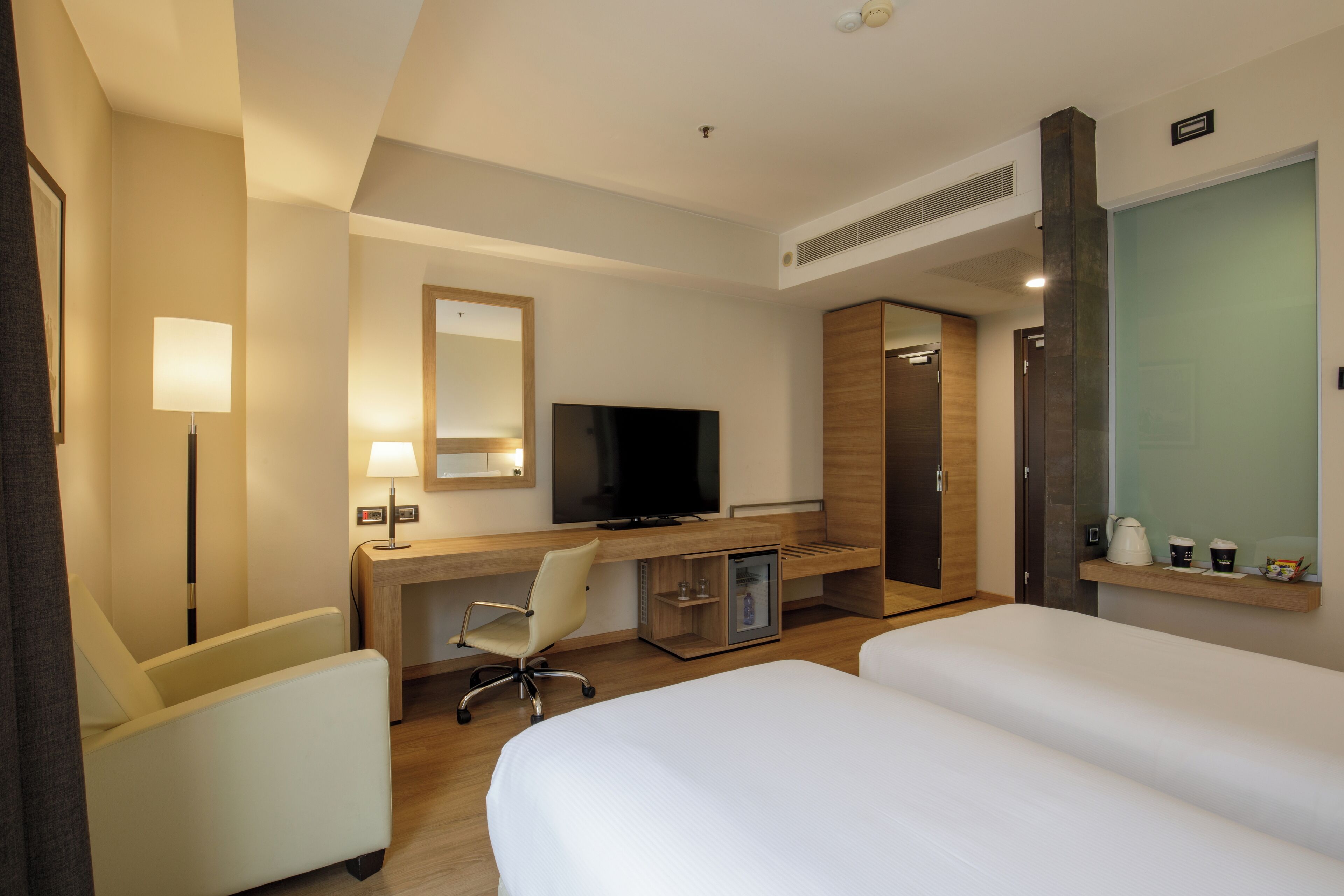 A hotel room with a bed, a desk with a computer, a TV, a chair, and a minibar.