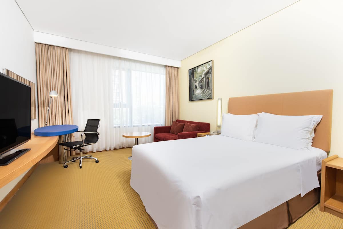 A hotel room with a large bed, a desk with a TV, a red armchair, a small table, and a painting on the wall.