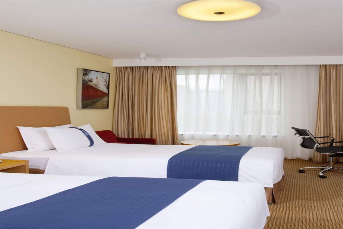 A hotel room with two beds, a desk, a chair, a lamp, and a large window with curtains.