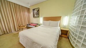 Habitación de hotel con una cama grande, una mesita de noche, una pequeña mesa con un frutero, una silla y una cortina.