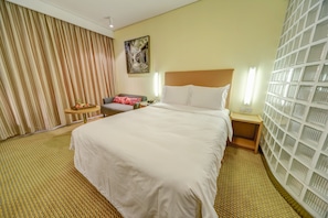 Quarto de hotel com uma cama grande, uma mesa de cabeceira, uma mesinha de apoio, uma pequena mesa com uma cesta de frutas, uma cadeira e uma cortina.