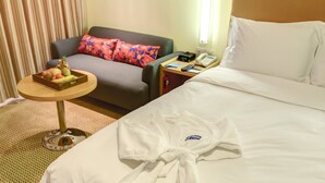 Habitación de hotel con cama, una mesita con frutero, sofá con cojines coloridos, un cuadro en la pared y una bata sobre la cama.