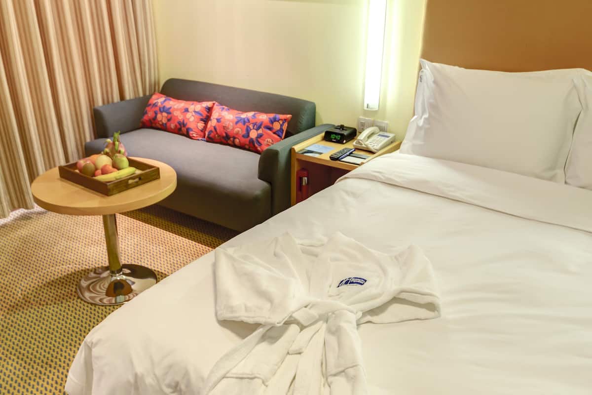 A hotel room with a bed, a small table with a fruit bowl, a sofa with colorful pillows, a painting on the wall, and a robe on the bed.