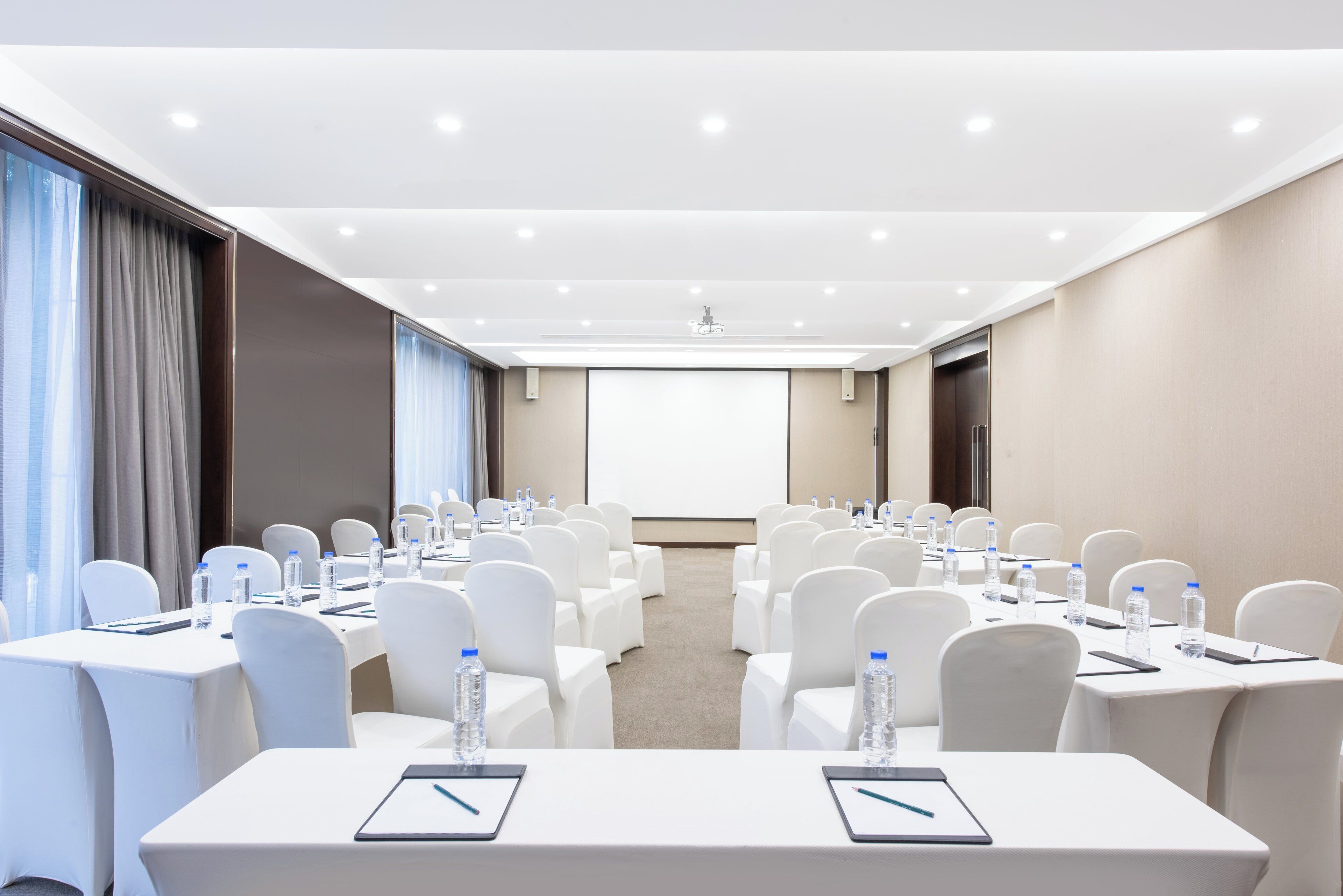 A conference room with rows of white chairs, tables with water bottles, and a projector screen at the front.