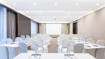 A conference room with rows of white chairs, tables with water bottles, and a projector screen at the front.