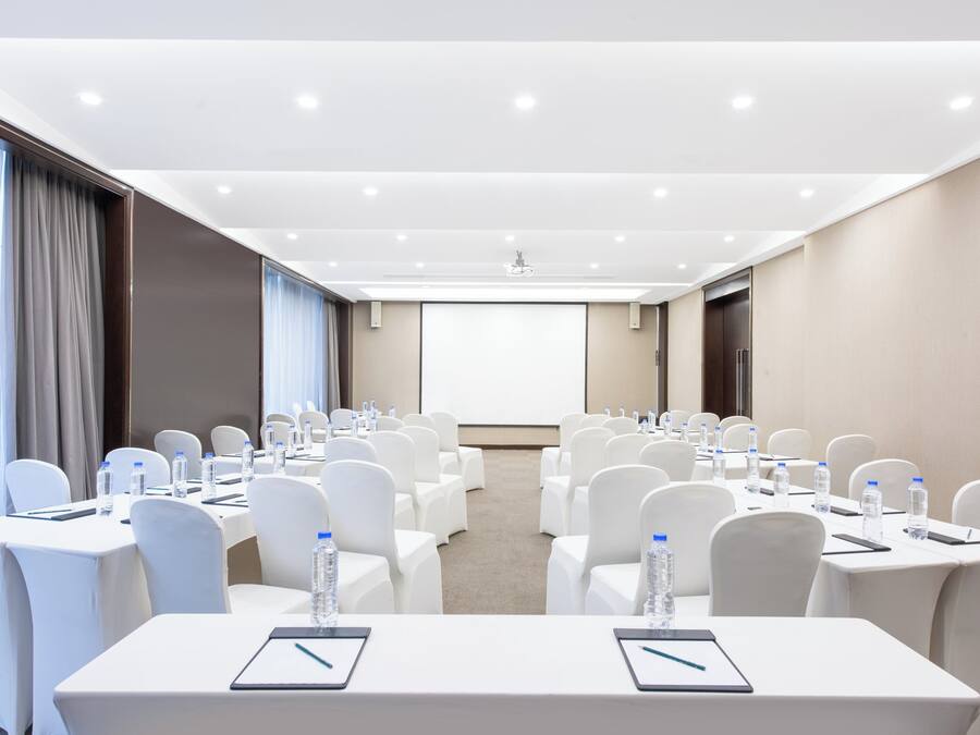 A conference room with rows of white chairs, tables with water bottles, and a projector screen at the front.