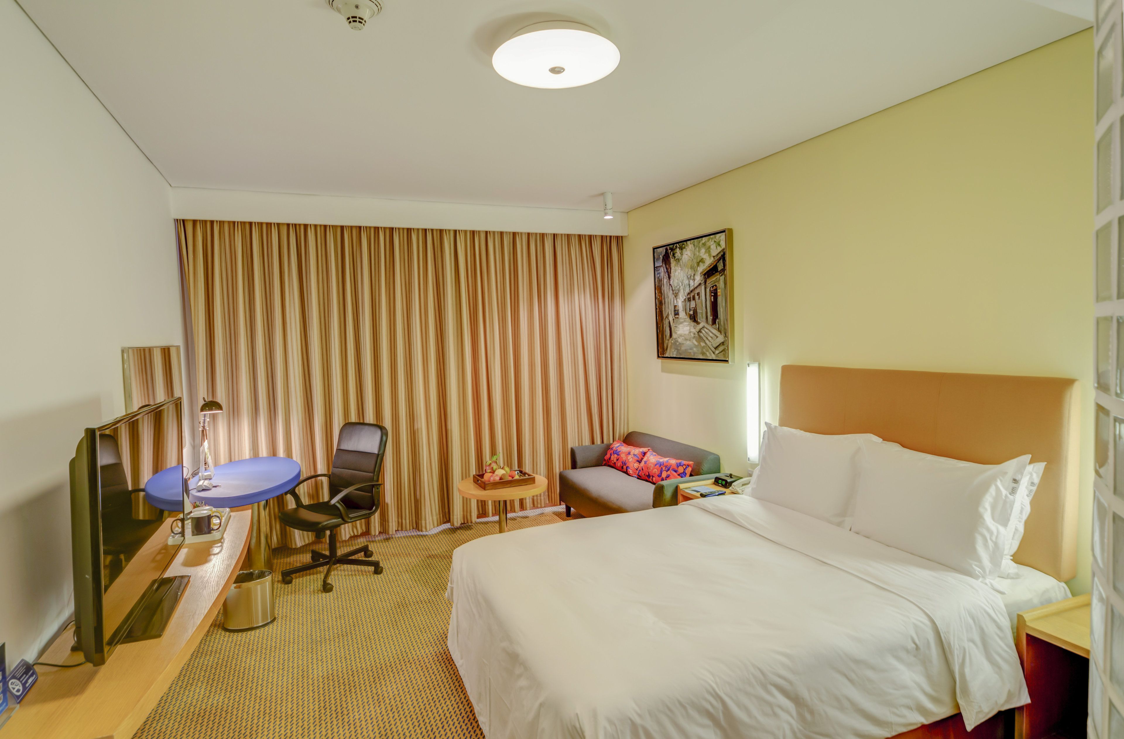 Una habitación de hotel con una cama, un escritorio con una computadora, una silla, una mesita con fruta y un cuadro en la pared.