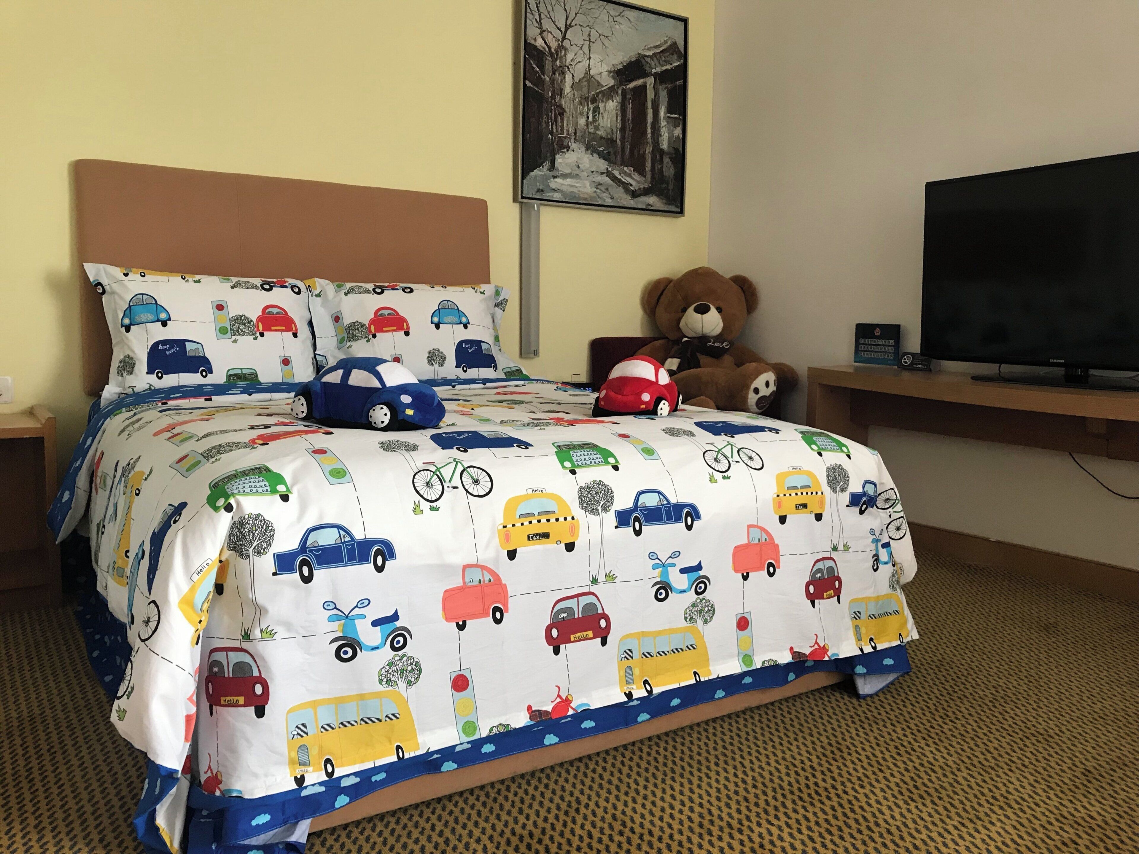 A bed with a vehicle-themed bedspread, a teddy bear, a television, and a painting on the wall.