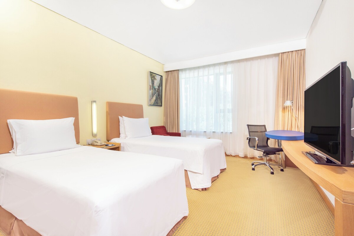 Quarto de hotel com duas camas, uma escrivaninha, uma televisão de tela plana, uma cadeira e uma janela com cortinas.