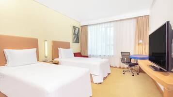 Quarto de hotel com duas camas, uma escrivaninha, uma televisão de tela plana, uma cadeira e uma janela com cortinas.