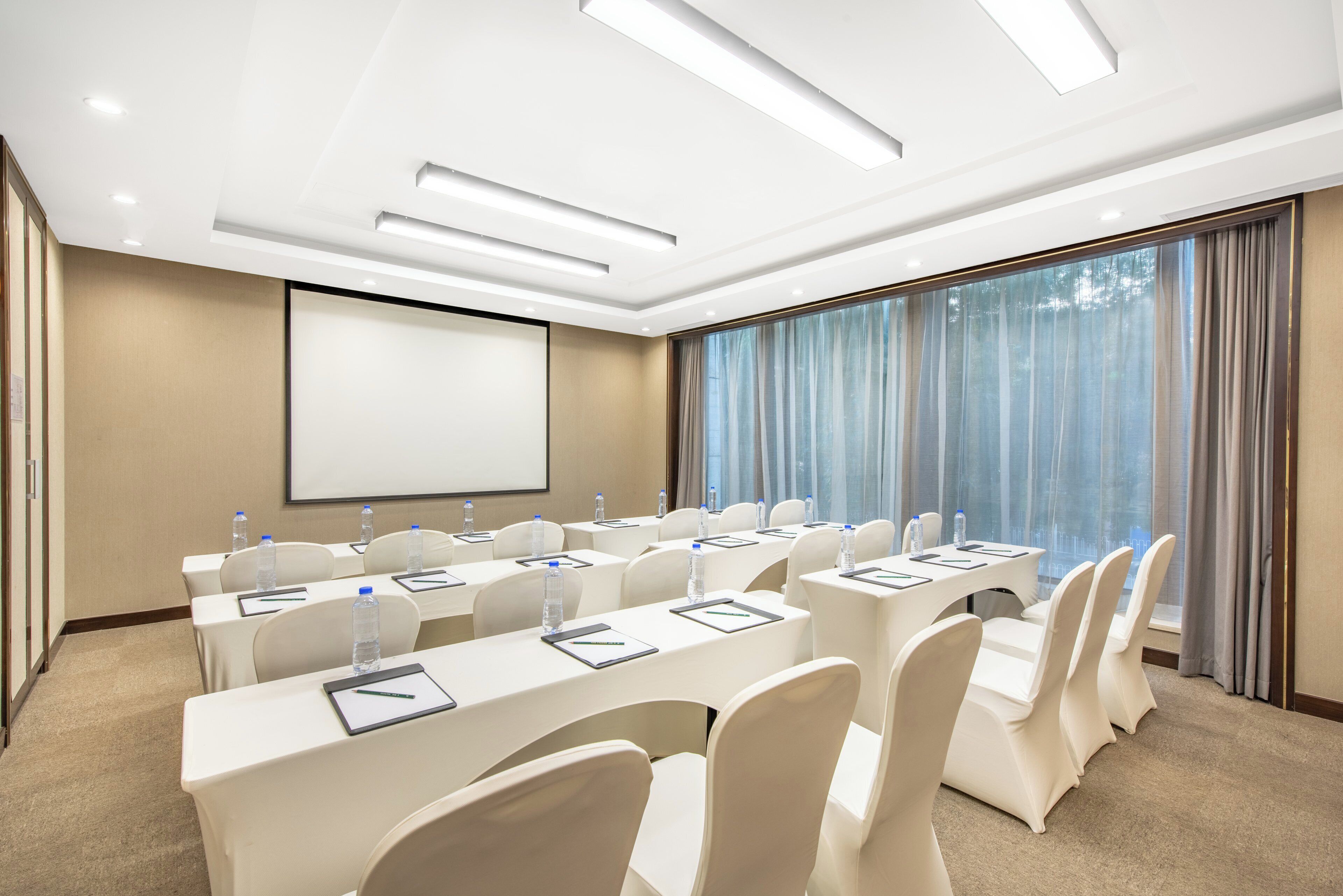 A conference room with a large screen, rows of tables with chairs, water bottles, and a view of trees through the window.