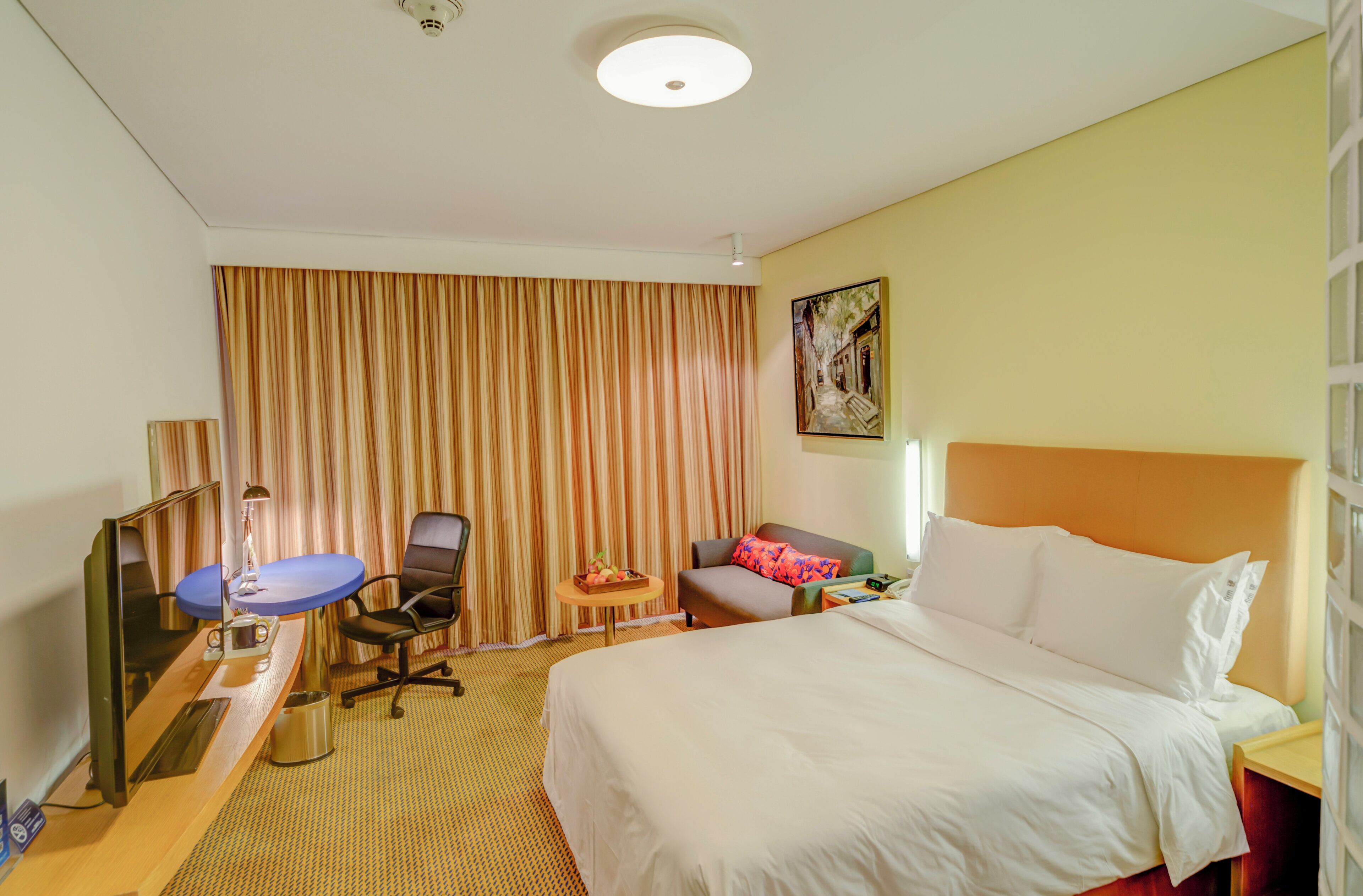 Habitación de hotel con cama, escritorio con computadora, una silla, una mesita con fruta y un cuadro en la pared.