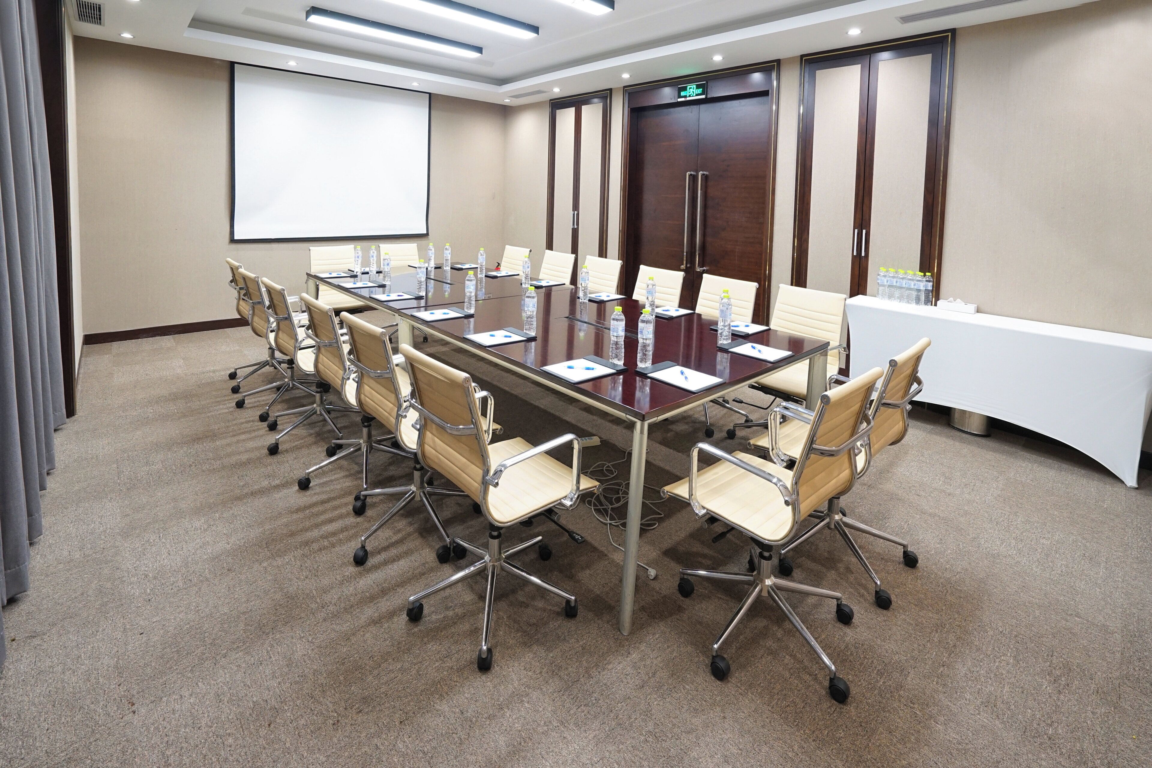 A conference room with a long table, chairs, a projector screen, and a door with an exit sign.