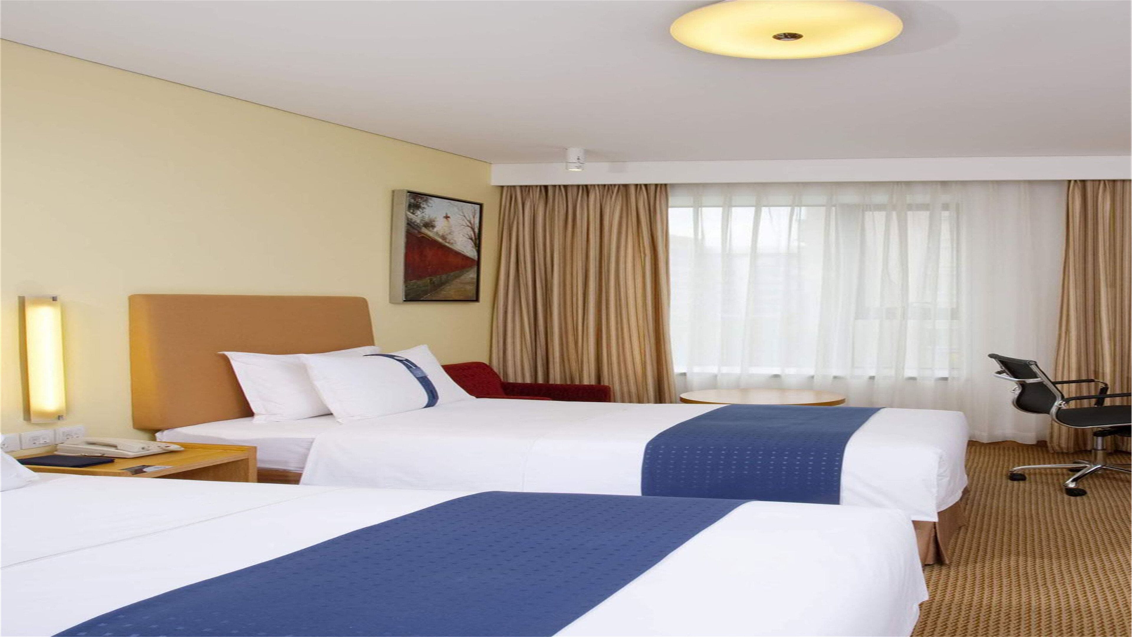 A hotel room with two beds, a desk, a chair, and a large window with curtains.