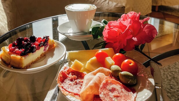A dining table with a cheesecake, fruit, and cured meats. A cup of coffee is on the table, and a vase with flowers is in the background. - Hotel Caroline (Brusimpiano)