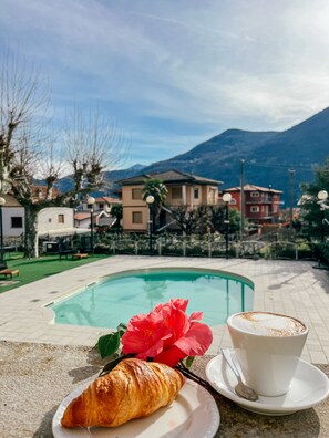 Ambiente a bordo piscina con un croissant, una tazza di caffè e un fiore rosso.