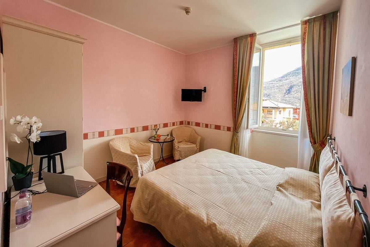 A hotel room with a bed, a desk with a laptop, a chair, a small table with a vase, and a window with curtains.
