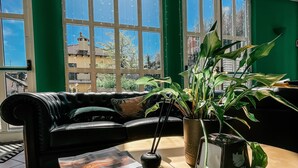 A room with a black leather sofa, a round table with a magazine, a bottle, and a plant. Large windows with green walls and a view of buildings are visible. - Hotel Caroline (Brusimpiano)